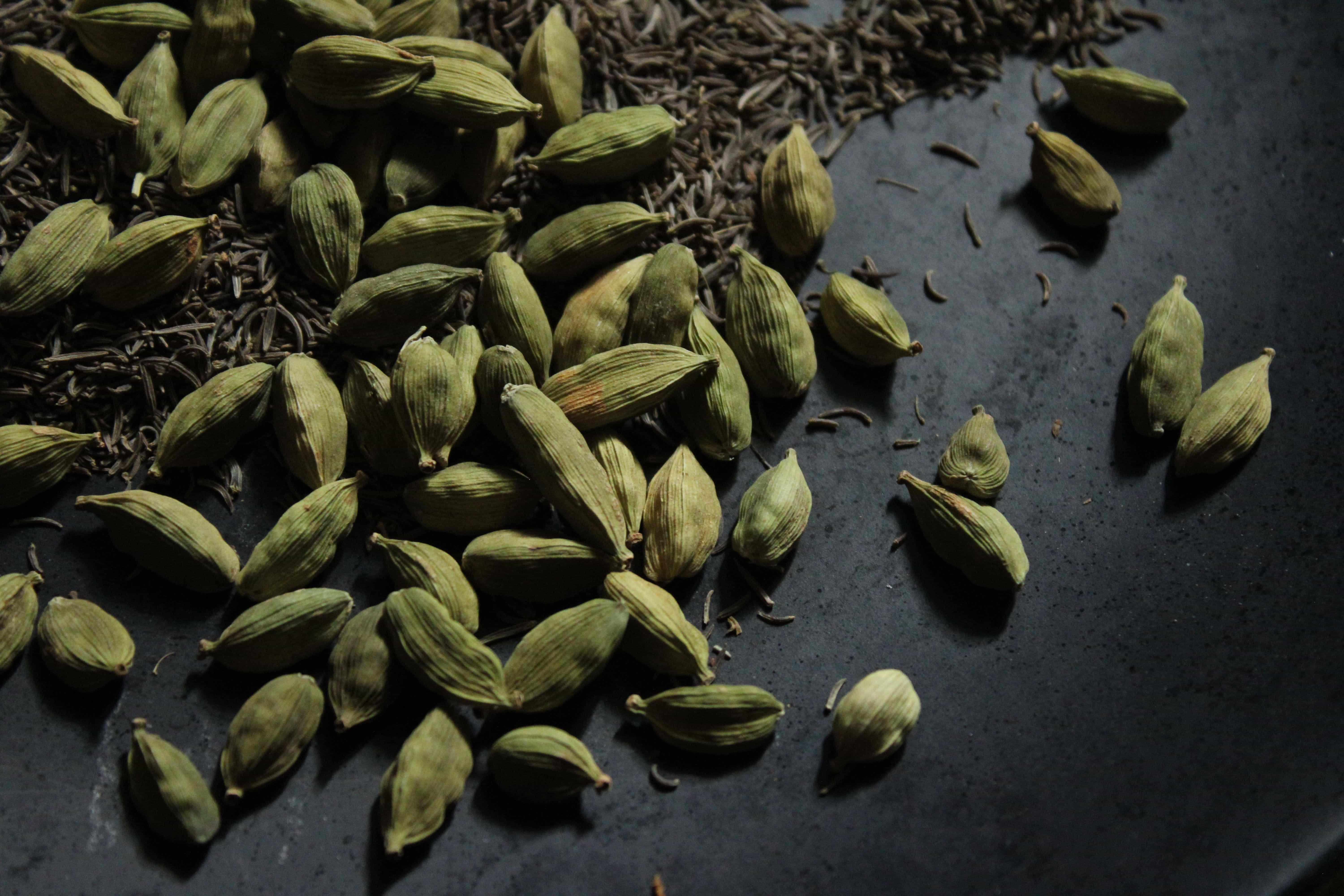Whole green cardamom pods scattered on a dark stone surface with cumin seeds.